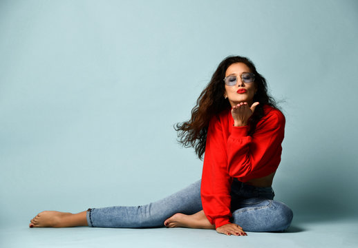 Curly brunette woman in smoked blue glasses, red sweater and blue jeans sits posing sends us a kiss 
