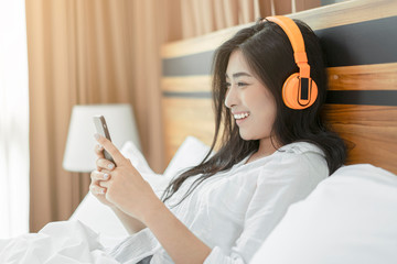 Asian beautiful woman resting in bed using touching mobile phone listening to music on wireless...