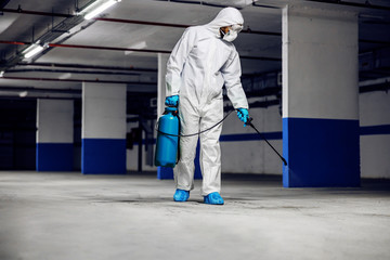 Full length of worker in protective sterile suit and mask disinfecting underground garage from corona virus / covid-19.