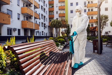 Full length of man in protective sterile suit disinfecting bench in park from corona virus. - obrazy, fototapety, plakaty