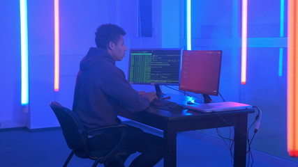 Young african man writing hacker programs in dark room with neon lights. He working on computer with two monitors.