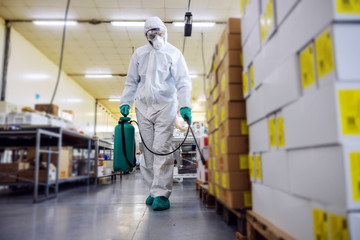 Man in protective suit and mask disinfecting warehouse full of food products from corona virus / covid-19. - obrazy, fototapety, plakaty