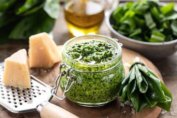 Wild leek pesto with olive oil and parmesan cheese in a glass jar on a wooden table. Useful properties of ramson. Leaves of fresh ramson. - obrazy, fototapety, plakaty