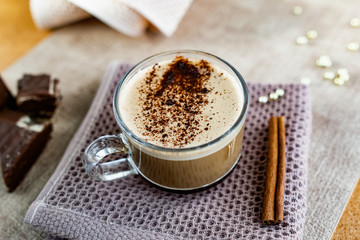 Hot cappuccino with chocolate chips on top of the foam, a cinnamon stick, Golden sugar beads and a sweet waffle next to it. Drink in a transparent Cup on a soft cloth