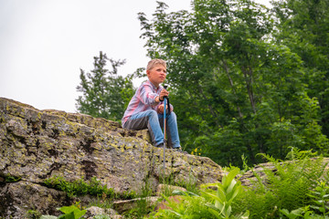 A boy traveler with a backpack and trekking poles is resting on a stone