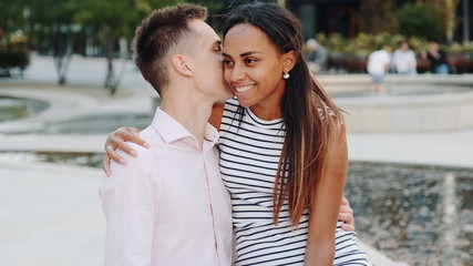 Close-up of young man whispering something in girlfriend's ear sitting on a bench outdoors. They smiling, hugging and talking to each other.