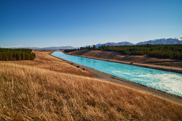 
Scenic view from a grassy hill overlooking a canal below with beautiful blue water flowing and glistening in the sunlight in Twizel in New Zealand South Island
