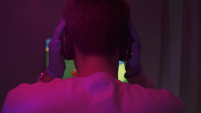 Young man in white sweater and medical gloves puts on his black headset and starts playing football video games at night with violet lights