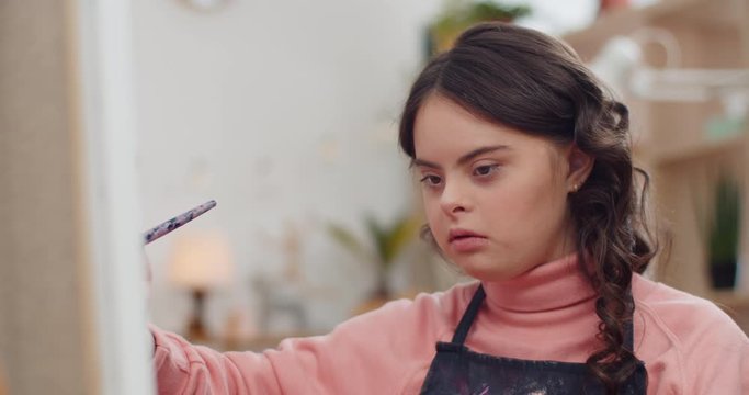 Close up view of talanted teen in aprone with down syndrome creating picture in her room.Beautiful girl sitting in front of molbert and painting. Concept of sunny children.