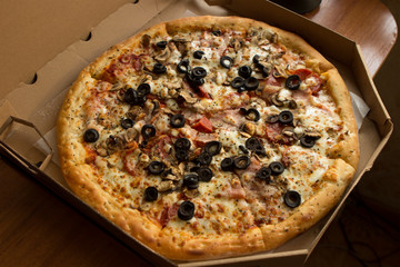 sliced pizza with black olives, champignon mushrooms, pepperoni salami, mozzarella cheese in a cardboard box. Pizza from home delivery is on the table in the apartment.