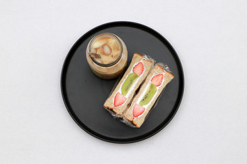 Breakfast in the black of round plate on the white floor. Strawberry and kiwi fruit with cream Sandwich, a glass of ice coffee.