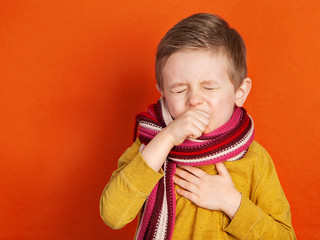 The European boy is ill, he coughs, covers his mouth with his hand. A thick scarf is wrapped around her neck. On an orange background. Treatment of colds and flu. Covid-19.