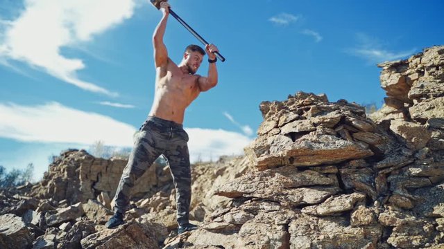 Muscular athlete with hammer on the rocks. Shirtless sportsman breaking stones with hard metal hammer outdoors. Slow motion.