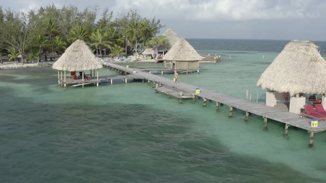 Aerial: Drone reversing from woman in bikini walking on pier by thatched roofs over sea - Coco Plum Cay, Belize
