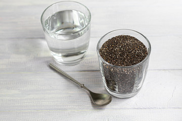 Black chia seeds in a glass bowl and water in which they are soaked. On a white background with a metal spoon.