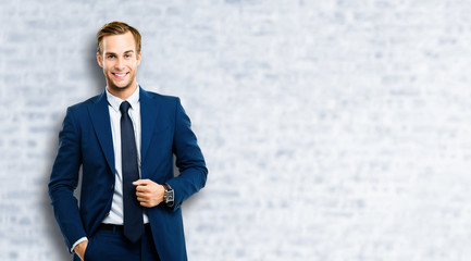 Portrait of happy confident businessman in blue suit and tie, standing against white brick wall...
