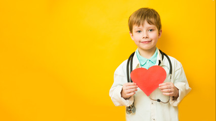 Cute child boy in doctor's uniform with stethoscope and heart in hand on yellow background. Copy...