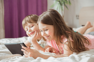 Happy children watching online movie stream with digital mobile tablet and lying on bed at home in morning, video call connection, social distancing, network technology concept