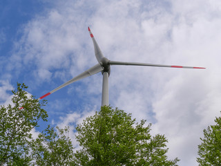 Windrad im Wald mit Blickpunkt nach oben nah am Windrad bei blauem Himmel mit weißen Wolken und hellgrünen Bäumen im Vordergrund