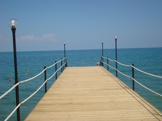 Fototapeta premium Wooden bridge with rope railing on the blue sea. Beautiful pier with lanterns for evening walks and diving. Blue sky and calm sea.