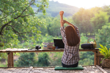Happy relaxed asian young woman sitting in the midst of nature with a laptop in the back of her stretching her arms above her head and looking out of the nature in the morning.