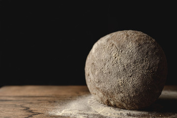 Close up of loaf of black whole meal cereal bread standing on a wooden table with flour. Black background and free space for text on the left.
