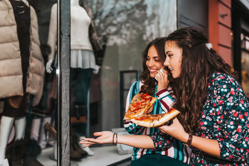 Portrait of two beautiful young women talking and eating pizza outdoors, and pointing finger in the shop window