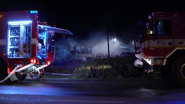 Firefighters try to put out a building on fire with hoses in an urban area at night - fire trucks in the foreground