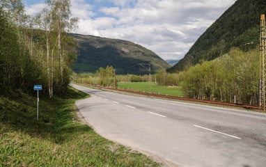 A bus stop on the Norwegian road. Railroad tracks. Green hills in the distance.