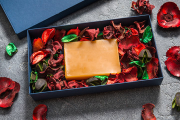 Bar of handmade soap in carton box with red potpourri flowers and leaves on black background, top view, mockup