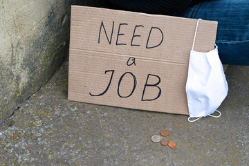 Unemployment concept and unemployed coronavirus sitting on the ground next to it a cardboard plate with the inscription need a job, the medical hygiene mask and coins