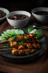vertical composition of chicken satay with slice of cucumber on stone plate and wooden tray