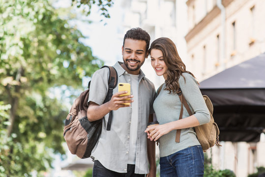 Young couple using smartphone outdoors. Joyful smiling woman and man looking at mobile phone in a city. Love, technology, connection, communication, summer travel concept