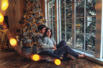 Couple of lovers married people on cozy sweaters hugging sitting under a Christmas tree in a house...