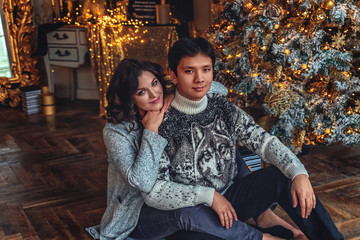 Couple of lovers married people on cozy sweaters hugging sitting under a Christmas tree in a house with a beautiful interior and a large window