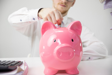Close up view of man in formal clothes putting coin to piggy bank, calculator on white desk. Savings, moneybox, thrift-box, saving, money banking, finance, commerce, payment, invest and buy concept