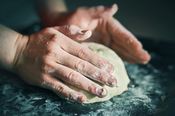 Fototapeta na wymiar The cook is kneading bread dough with strong hands on a dark baking tray strewn with white crumbly flour, illuminated by light.
