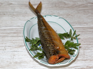 Smoked mackerel on decorative plate, few green herbst around, on wooden table close up
