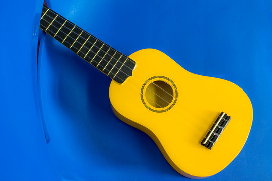 Yellow Ukulele. Child Uke On A Blue Plastic Kids Chair Background.