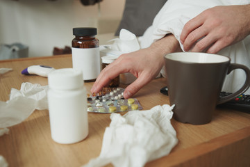 Hands of sick man taking tablets from table while sitting on bed or couch