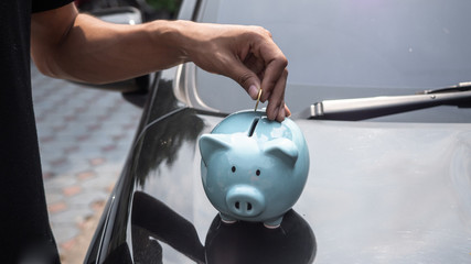 A young man is standing frount of a car and putting money into piggy bank. Saving to buy a house, home savings, loans market concept.