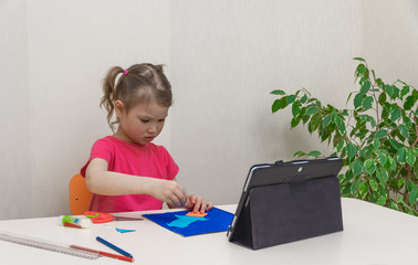 Distance learning online education. A preschool girl with a digital tablet looks at it and doing homework. Paper, stationery on table. Application of colored paper