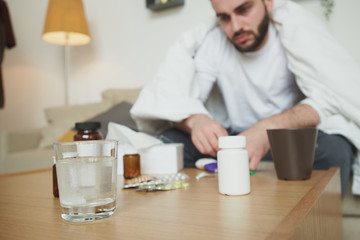 Sad and sick young man wrapped in blanket sitting in front of table with pills