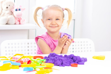 Obraz na płótnie Canvas happy Girl plays kinetic sand in quarantine. Blond beautiful girl smiles and plays with purple sand on a white table.