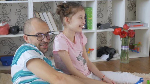 The father and daughter play together on the floor with the constructor, while the eldest daughter sits on the sofa and uses a smartphone.