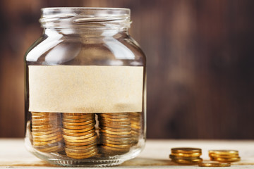 A glass jar with coins and a sticker with free space for text, on a wooden table.