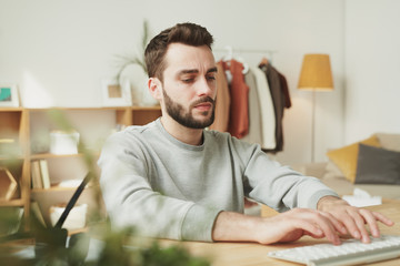 Obraz premium Serious young businessman in grey sweatshirt typing in front of laptop