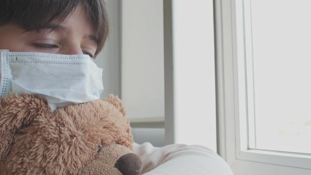 boy in a medical mask with a soft toy sits near the window and looks longingly at the street