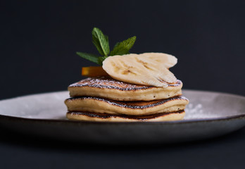 Homemade breakfast of pancakes decorated with orange, banana, mint and sugar powder on black background in grey plate.
