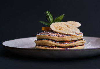 Homemade breakfast of pancakes decorated with orange, banana, mint and sugar powder on black background in grey plate.